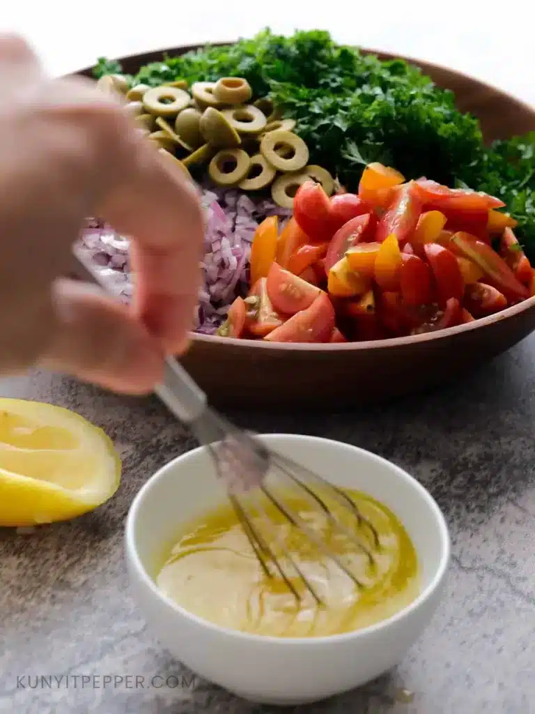 Whisking Dressing for Parsley Tomato Salad in a small bowl with a big bowl of chopped vegetables behind it