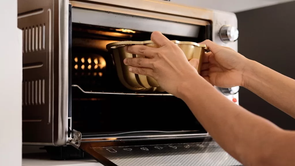 Step 6 on how to make gingerbread bundt cake. photo shows my hand putting the cake pan in the oven