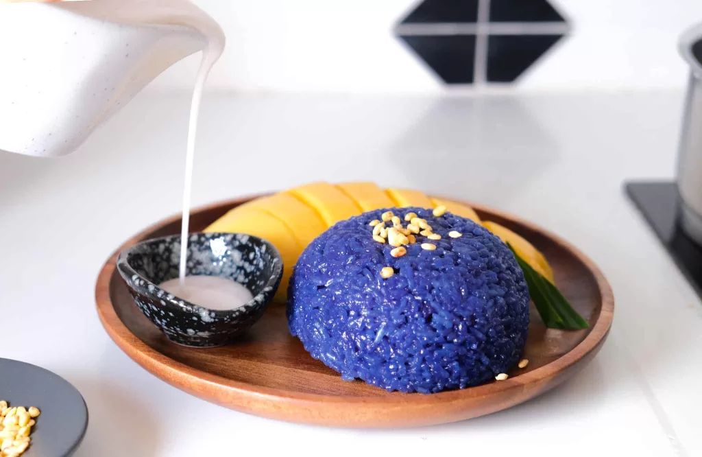 Pouring coconut milk into a small bowl next to sliced mango and butterfly pea sticky rice